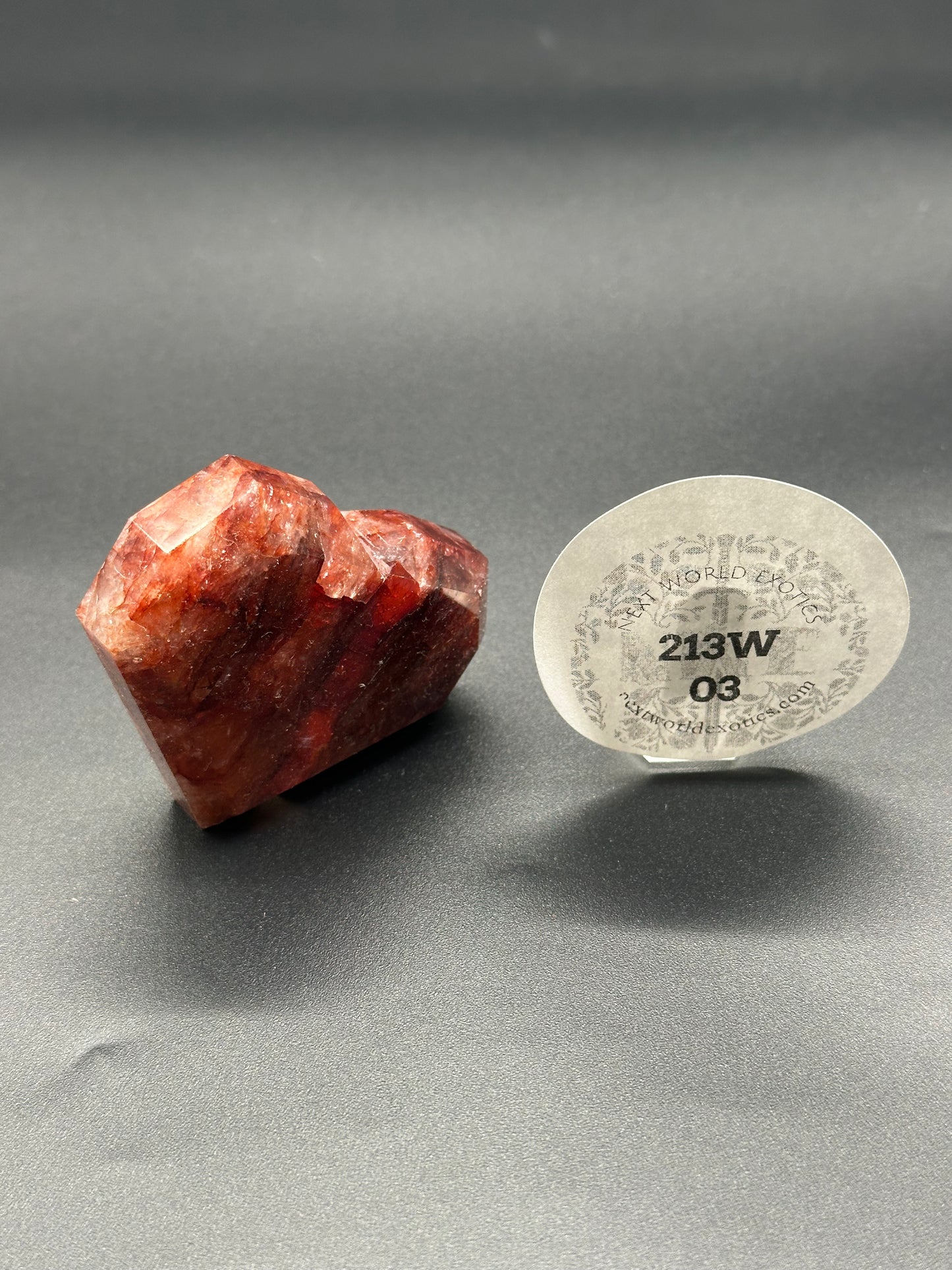 Fire Quartz Faceted Heart (213W03) displayed on a black surface, featuring a distinct faceted design, accompanied by a nearby label detailing product specifics.