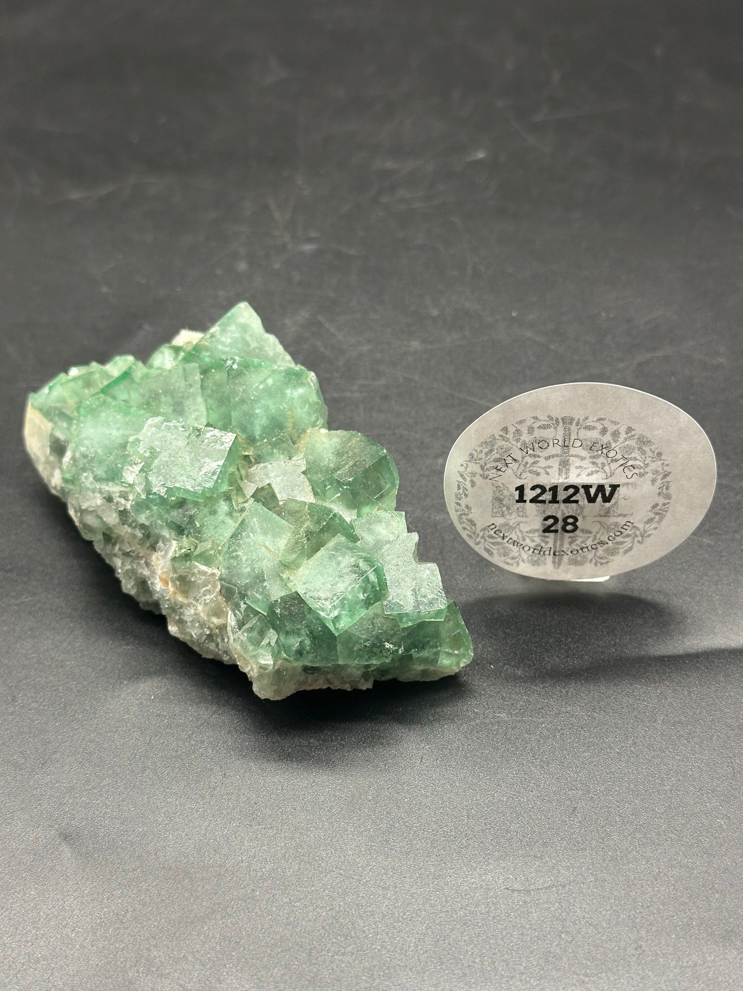 Raw Heliofluorite Carving (1212W28) featuring a green crystal with a round label, displayed on a dark surface.