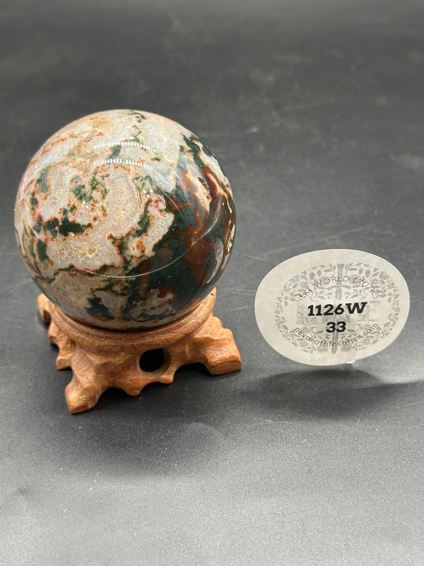 Red Moss Agate Sphere (1126W33) displayed on a wooden stand, accompanied by a paper tag.