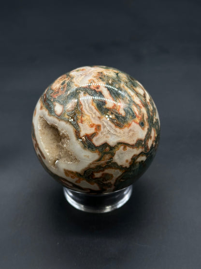 Red Moss Agate Sphere (812W01) with swirling bands and a geode cavity filled with druzy crystals, displayed on a clear stand.