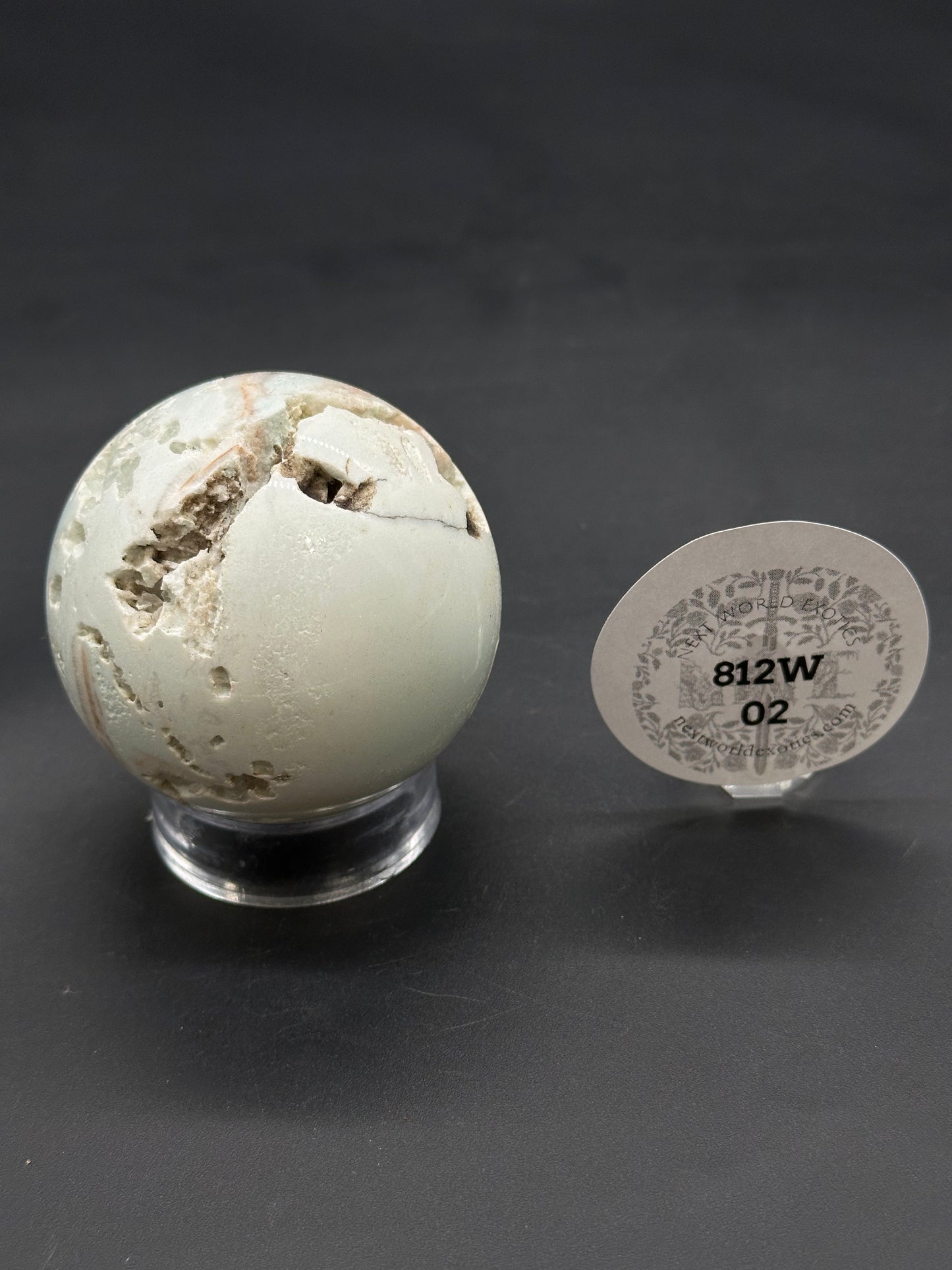 Caribbean Calcite Sphere (812W02) featuring natural cavities and brown veins, displayed on a clear acrylic stand, with an oval plaque beside it.