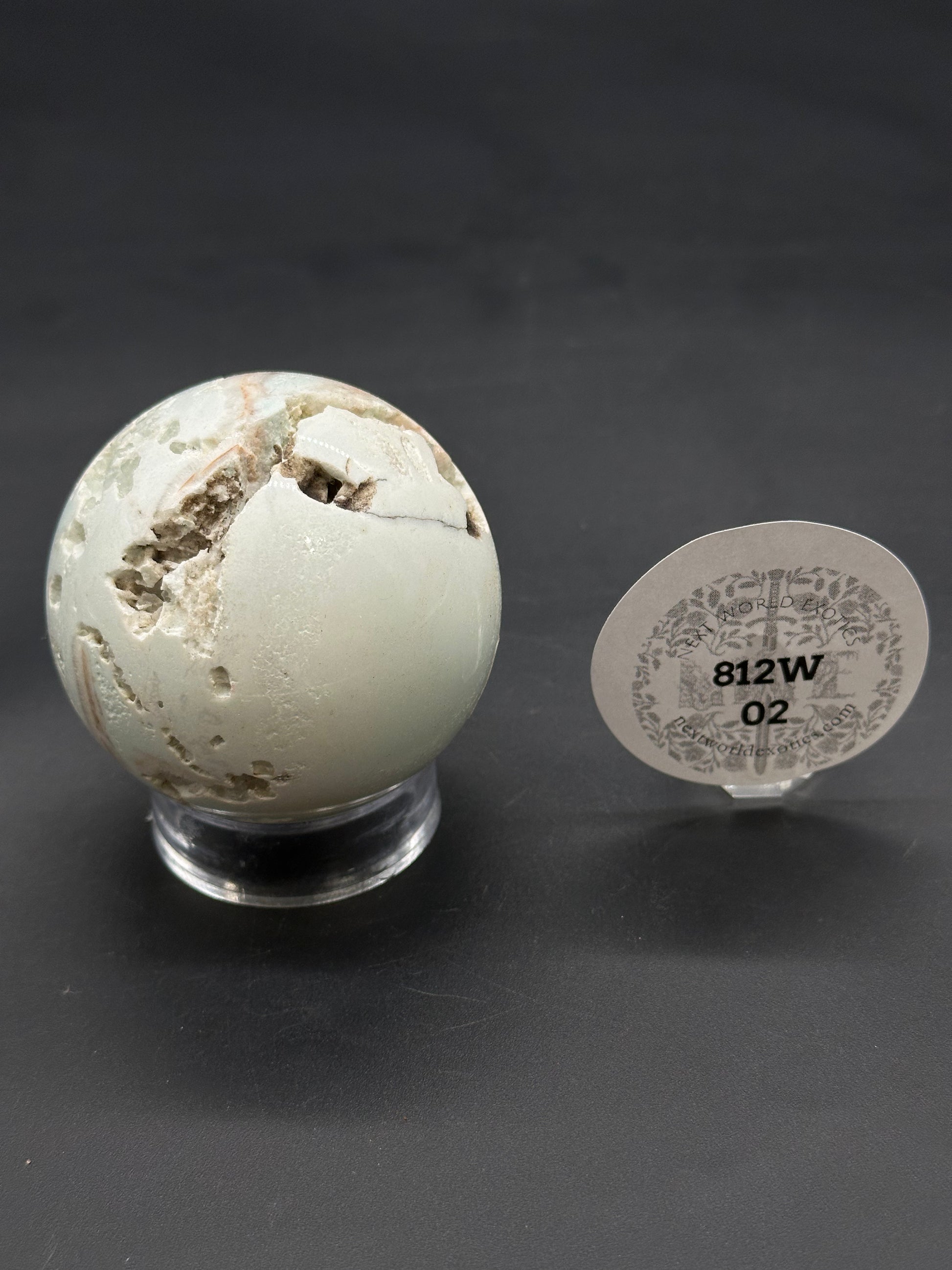 Caribbean Calcite Sphere (812W02) featuring natural cavities and brown veins, displayed on a clear acrylic stand, with an oval plaque beside it.