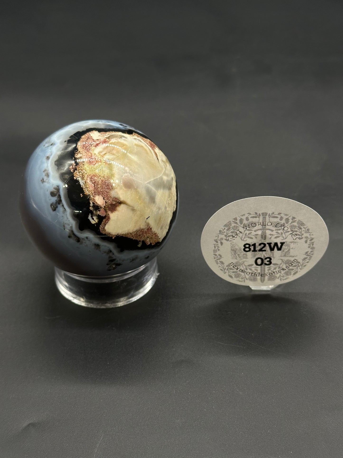 Volcanic Agate Sphere (812W03) on acrylic stand, showcasing mottled swirls; resembles ocean jasper. Circular label “812W 03” from “New World Exotics” nearby.