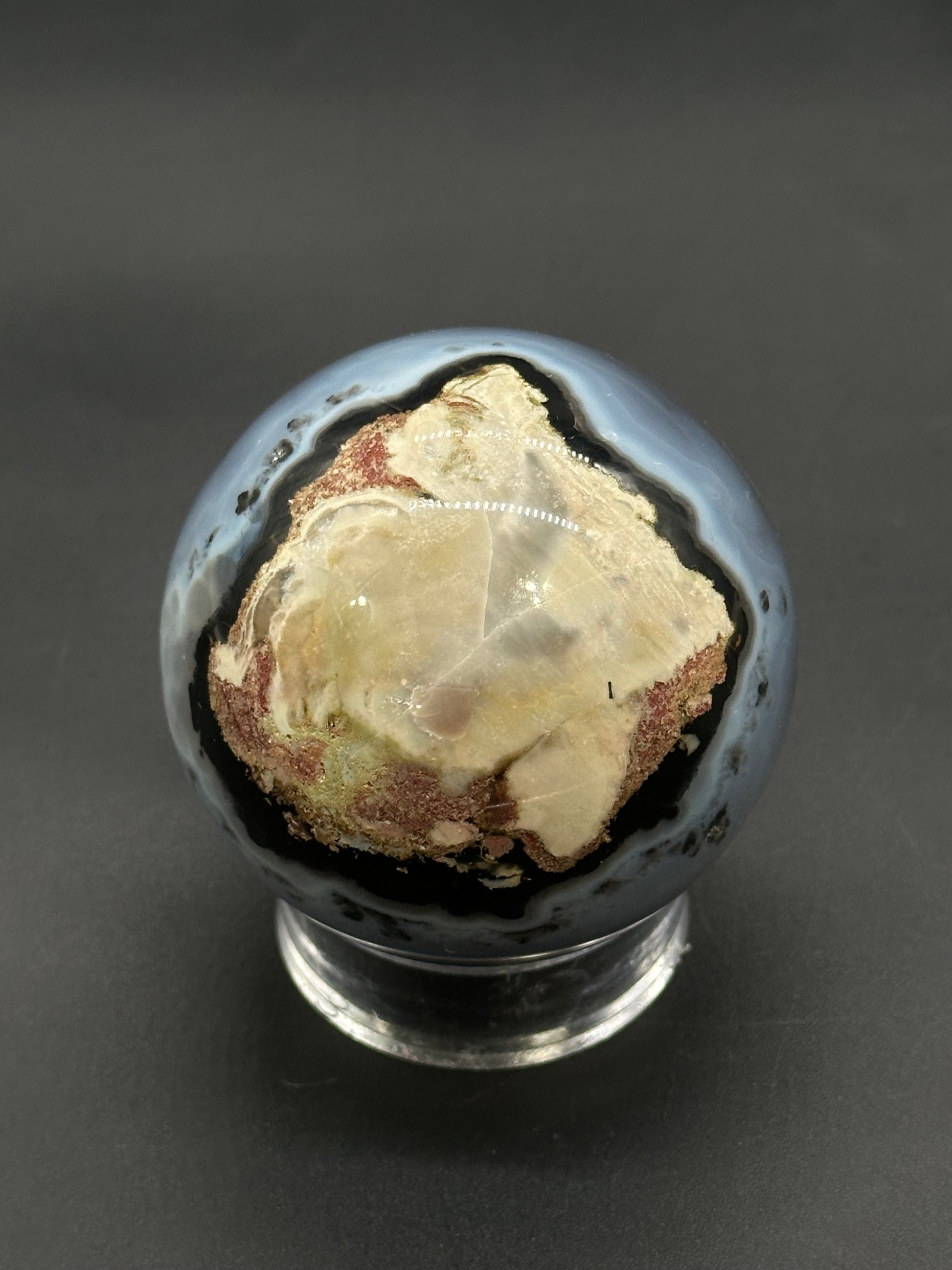 Volcanic Agate Sphere (812W03) displayed on a clear stand, showcasing its banded rim and crystalline center, highlighting its crafted mineral beauty.