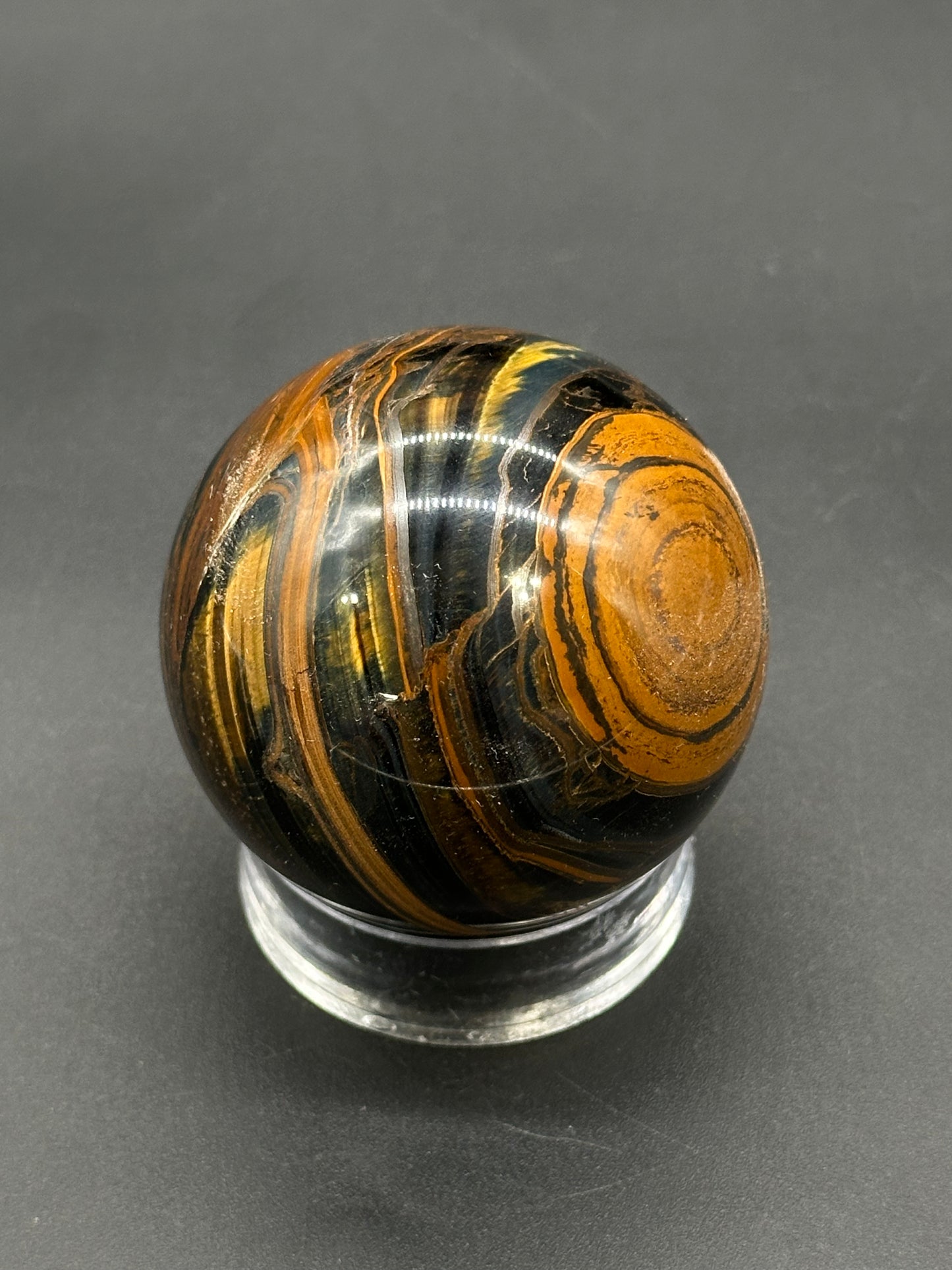 Tigers Iron Sphere (812W04) on a clear stand, showcasing its polished, swirling layers of black, golden-brown, and rust-orange, typical of tiger iron minerals.