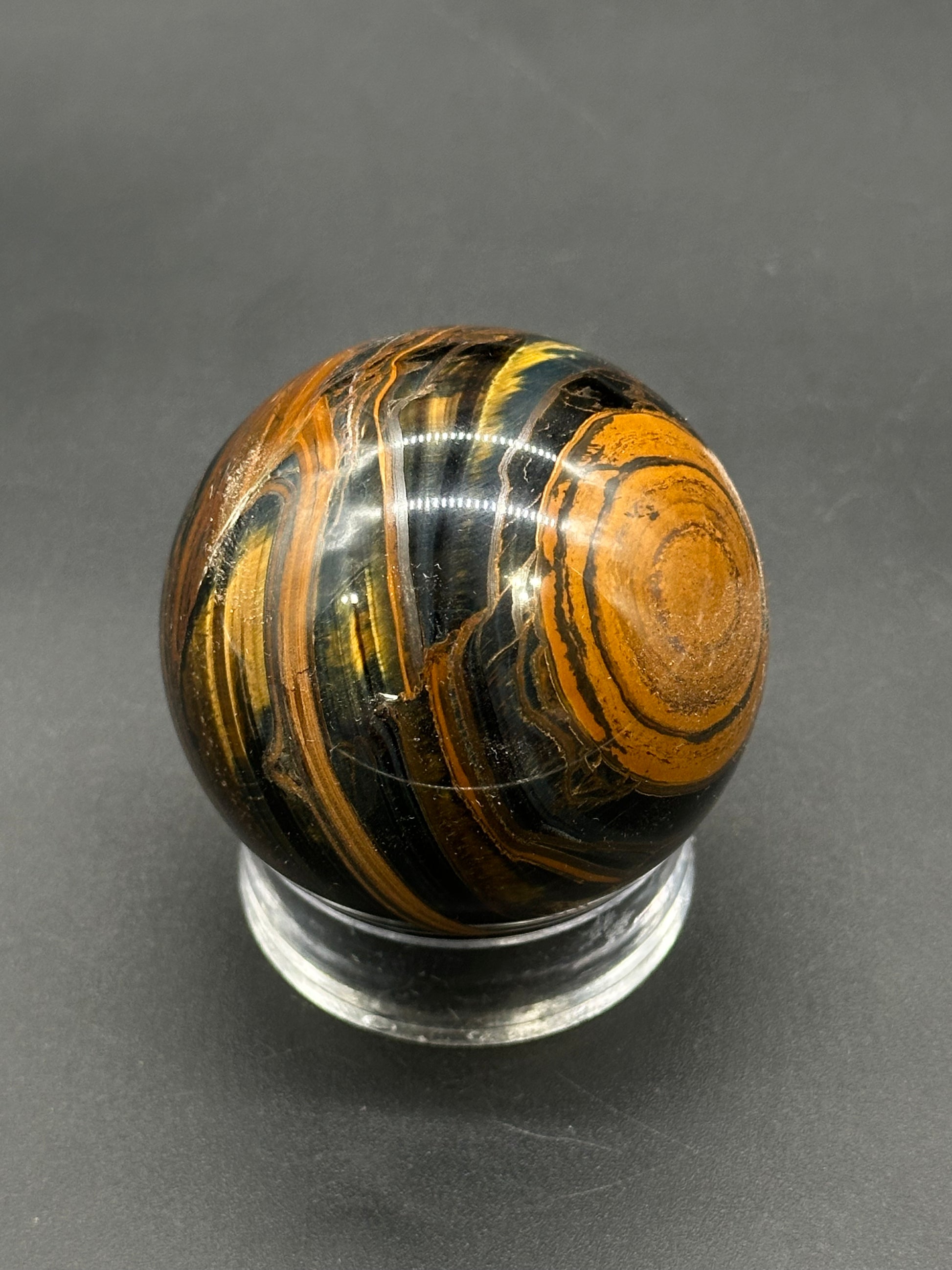 Tigers Iron Sphere (812W04) on a clear stand, showcasing its polished, swirling layers of black, golden-brown, and rust-orange, typical of tiger iron minerals.