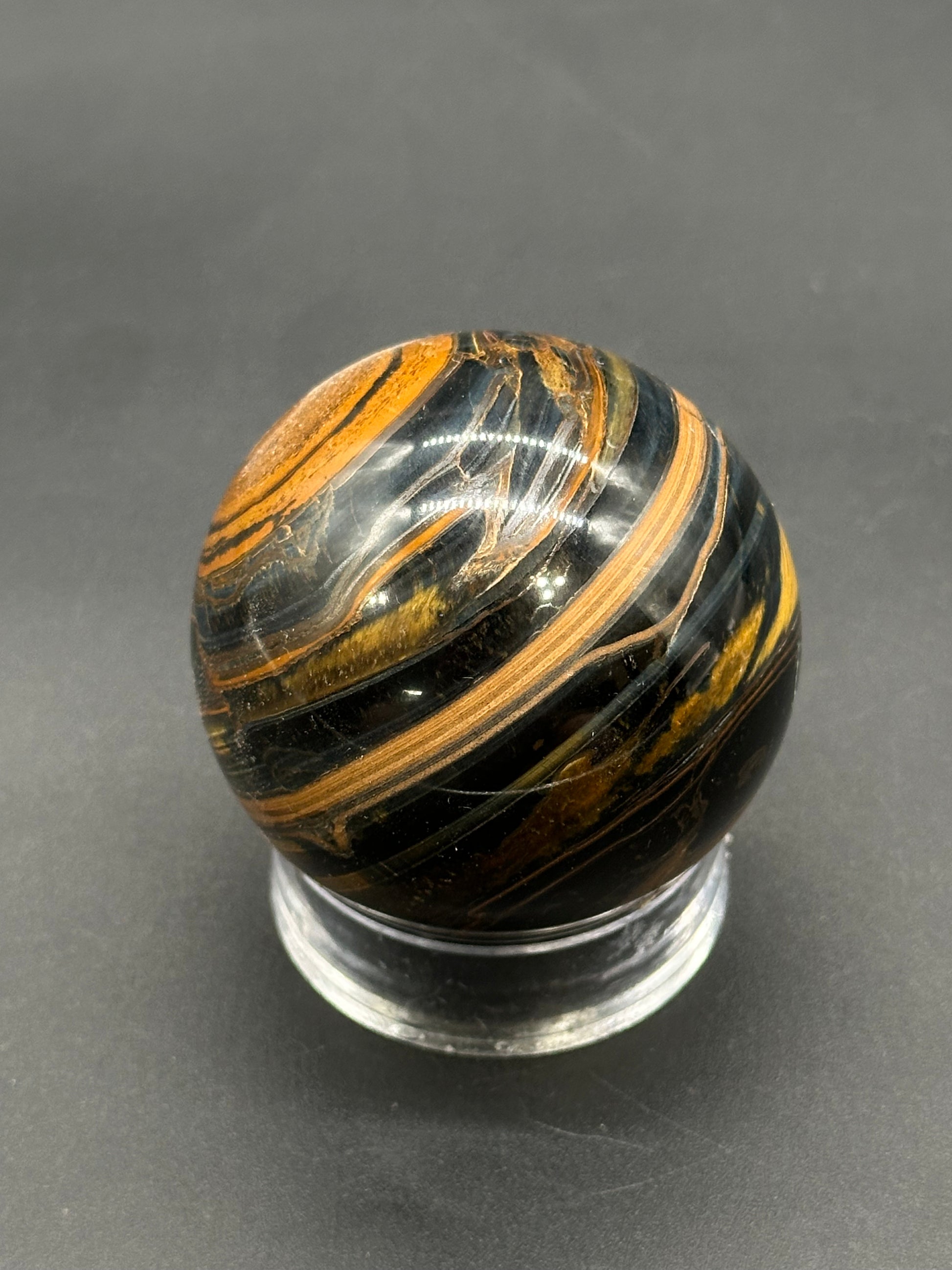 Tigers Iron Sphere (812W04) features glossy black and brown-gold swirls, resting on a clear acrylic stand, ideal for decorative or collectible use.