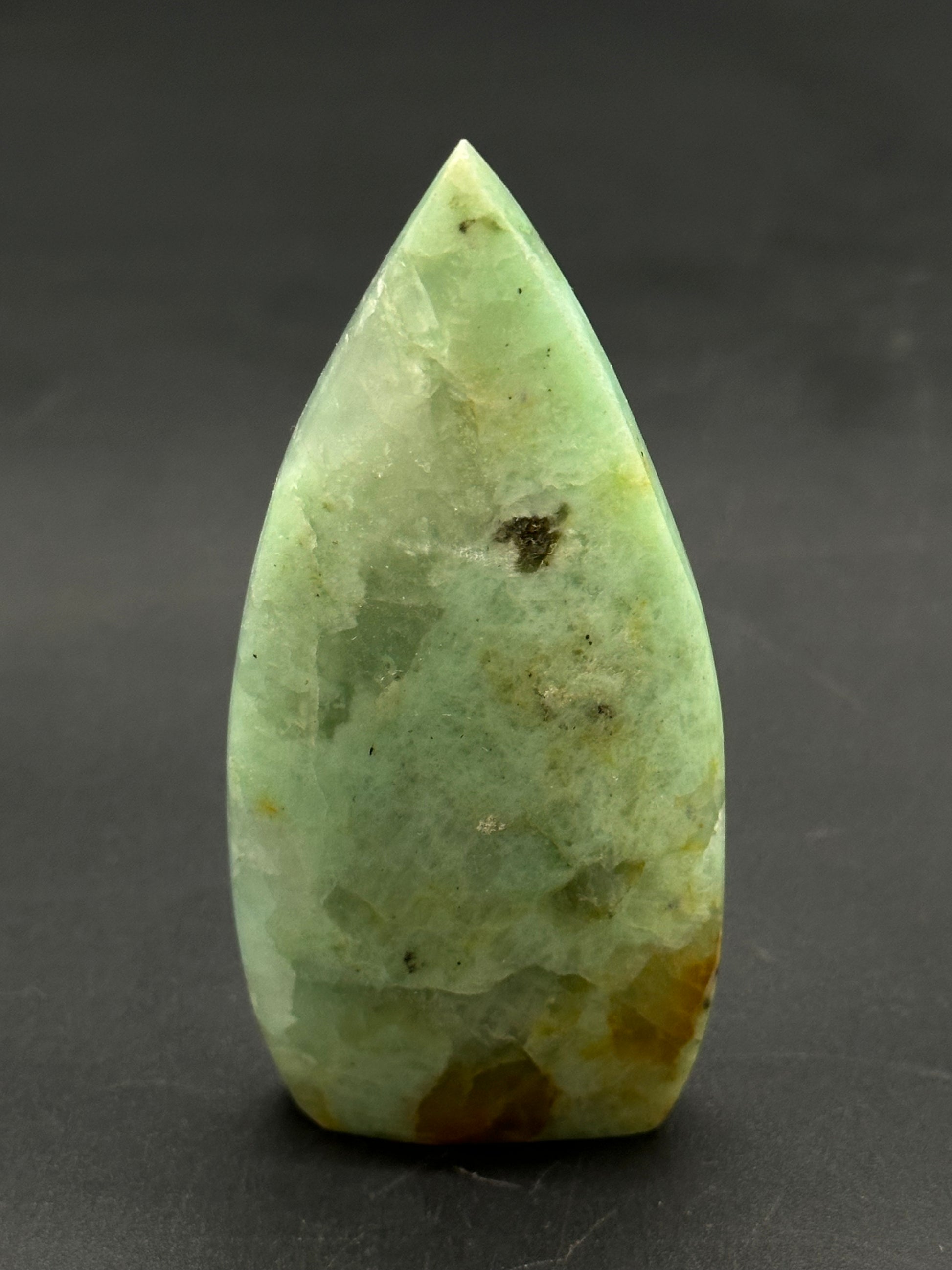 Quantum Quatro Free Form (812W07) polished green stone with brown inclusions, shaped like a tapered flame-tip, displayed upright on a dark surface.