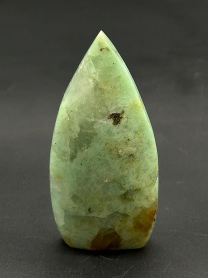 Quantum Quatro Free Form (812W07) polished green stone with brown inclusions, shaped like a tapered flame-tip, displayed upright on a dark surface.