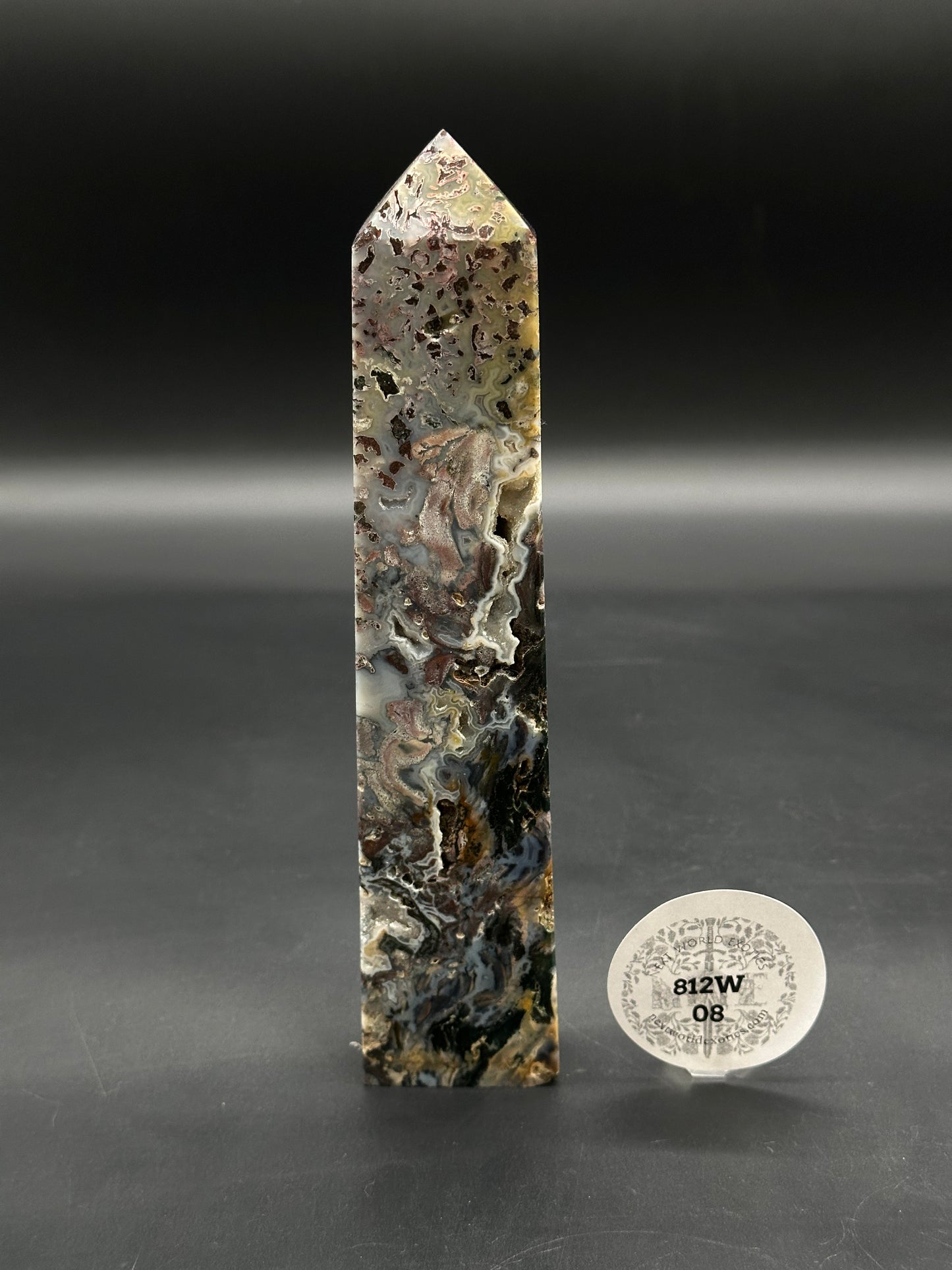 Ocean Jasper Tower (812W08) features a polished obelisk with swirling moss-like patterns in grays and browns, mounted upright, accompanied by a small label on a clear stand.