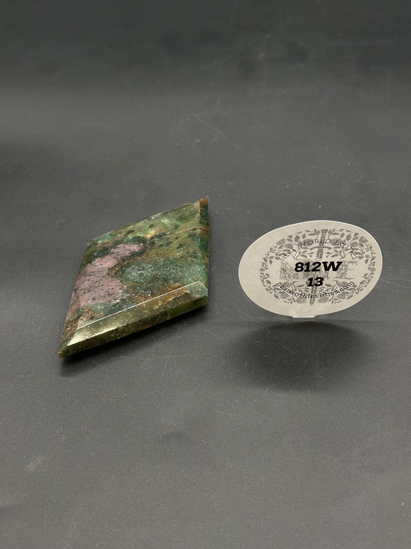 Ruby Kyanite Faceted Diamond (812W13) on a dark surface, skewed diamond shape with beveled edges, accompanied by a clear stand holding a labeled tag.
