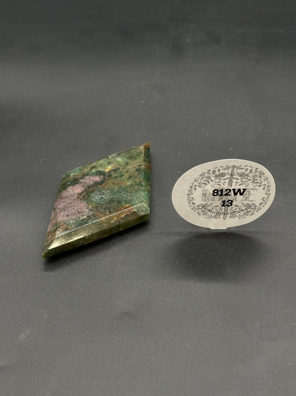 Ruby Kyanite Faceted Diamond (812W13) on a dark surface, skewed diamond shape with beveled edges, accompanied by a clear stand holding a labeled tag.
