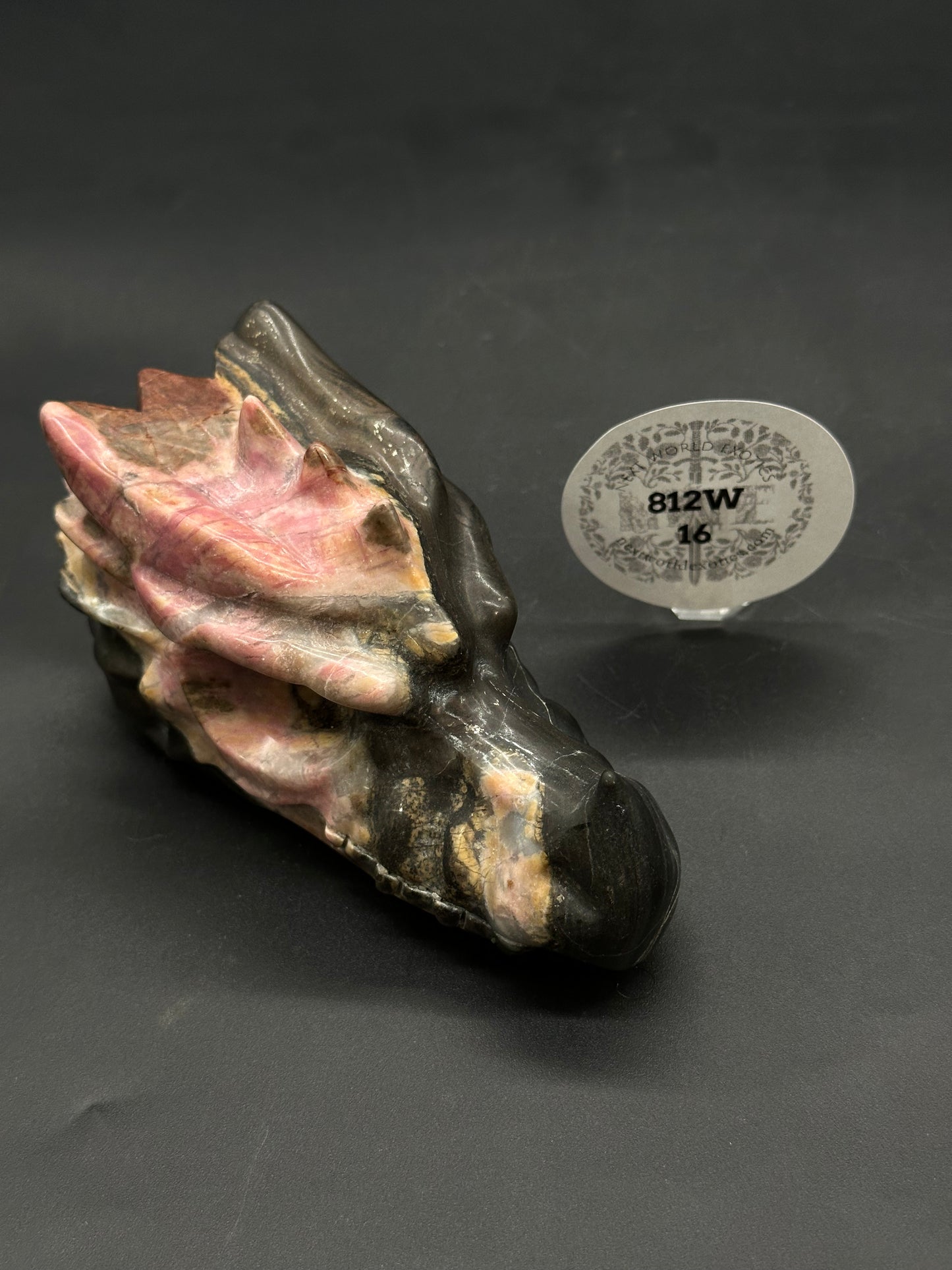 Rhodonite Dragon Carving (812W16) showcasing intricate detailing in banded stone, displayed on a black surface with a clear stand and round label nearby.