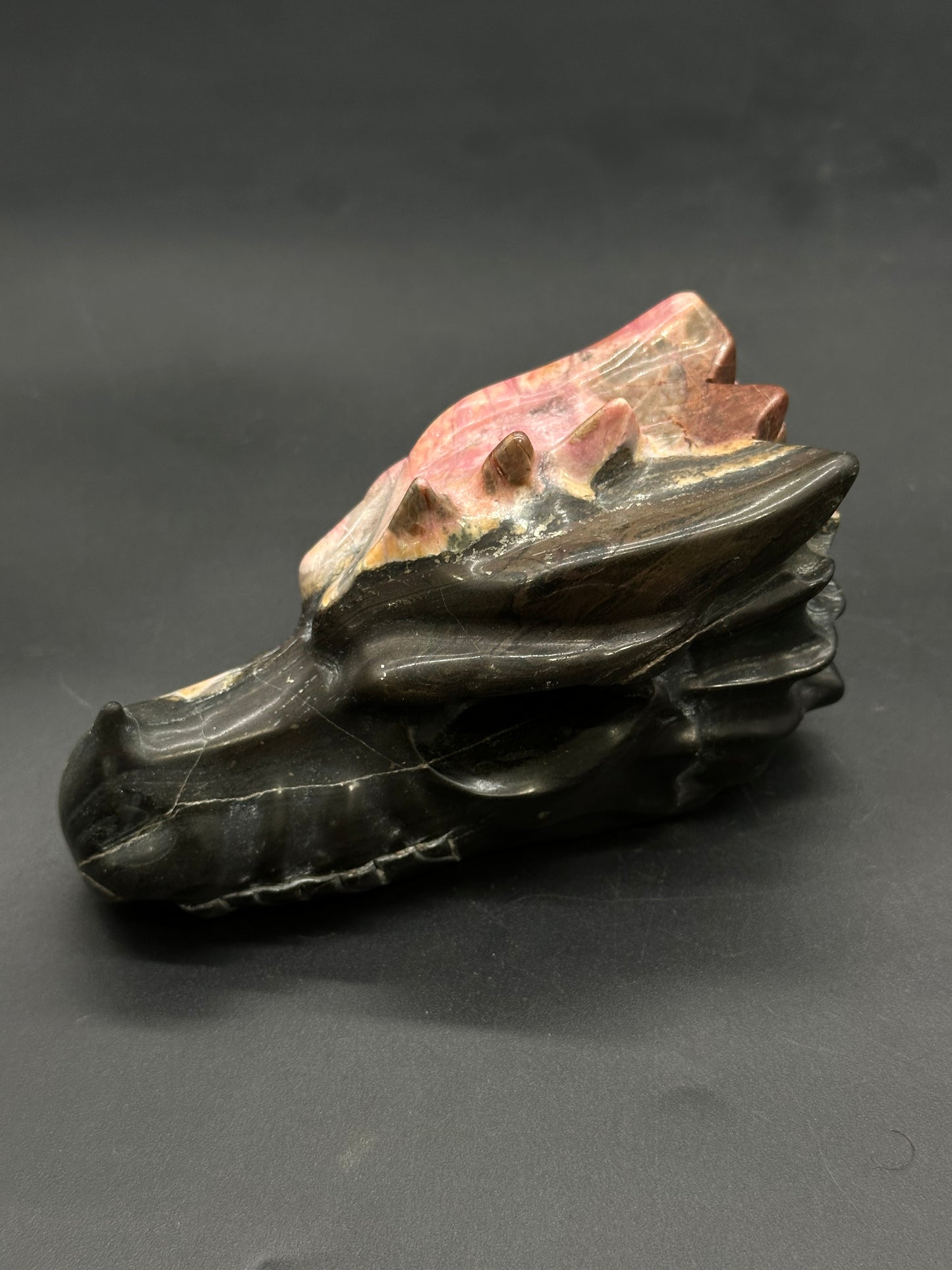Rhodonite Dragon Carving (812W16) features a hand-carved stone skull with polished eye sockets, teeth, and ridged horns, showcasing natural banding for a dragon-like appearance.