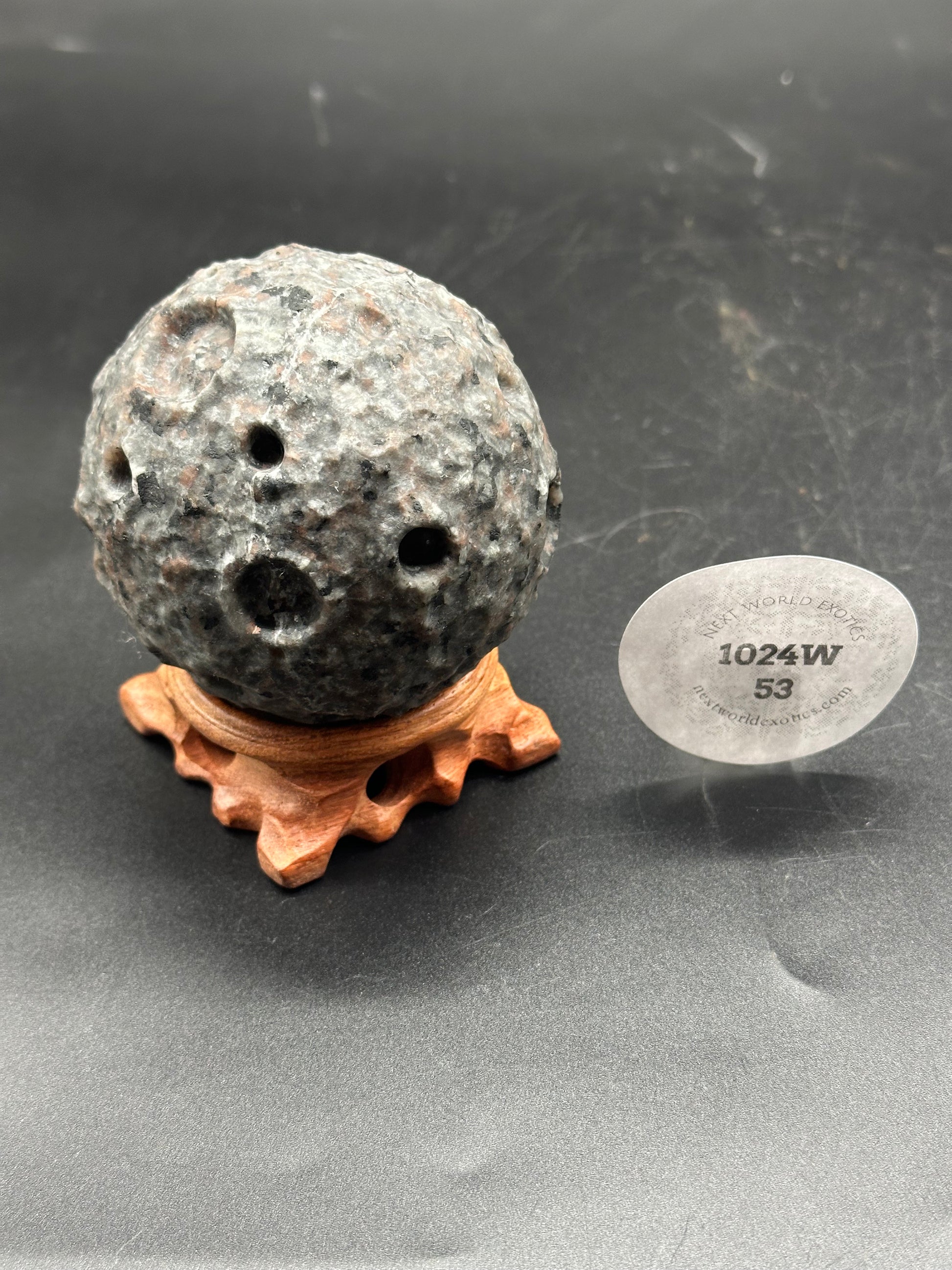 Yooperlite Moon Carving (1024W53) featuring a round stone ball with natural holes, displayed on a wooden stand, showcasing its unique mineral texture.