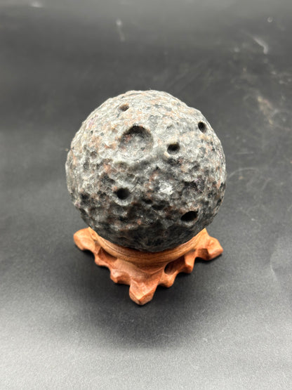 Yooperlite Moon Carving (1024W53) on a wooden stand, featuring a distinct round shape with a visible hole, highlighting its unique mineral structure.