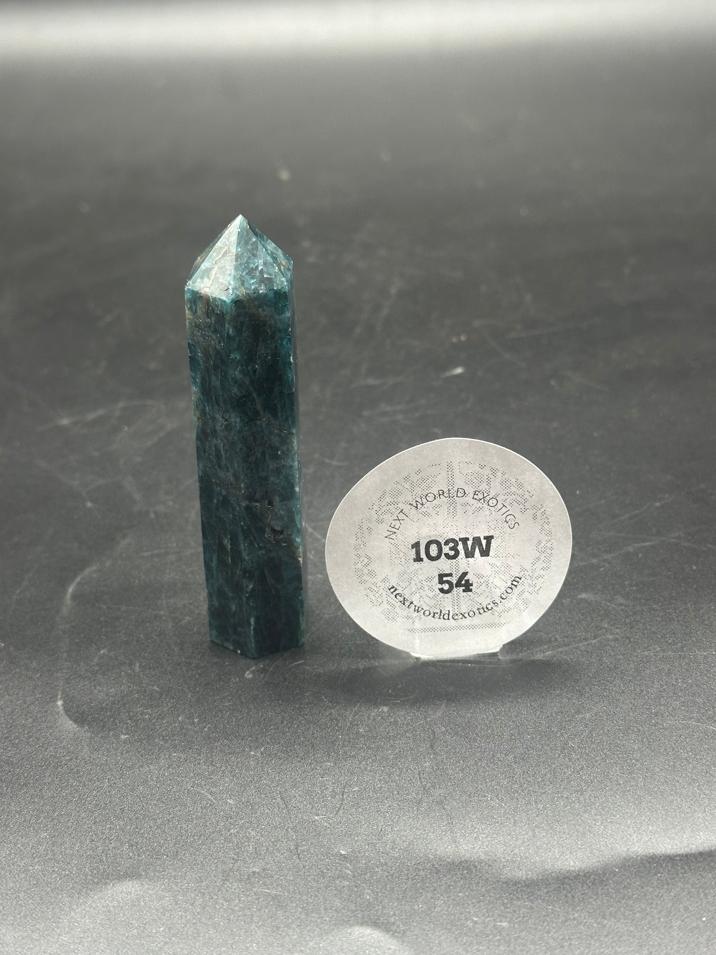 Blue Apatite Tower (103W48) featuring a polished crystal with intricate patterns, standing upright, accompanied by a label with black text and numbers in the background.