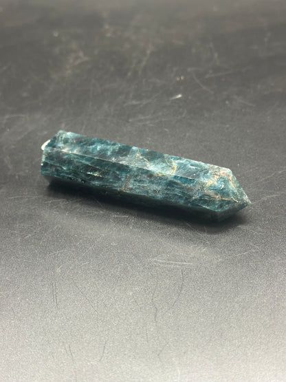 Blue Apatite Tower (103W48), a polished crystal with pointed edges, displayed on a dark surface, highlighting its natural structure and mineral composition.