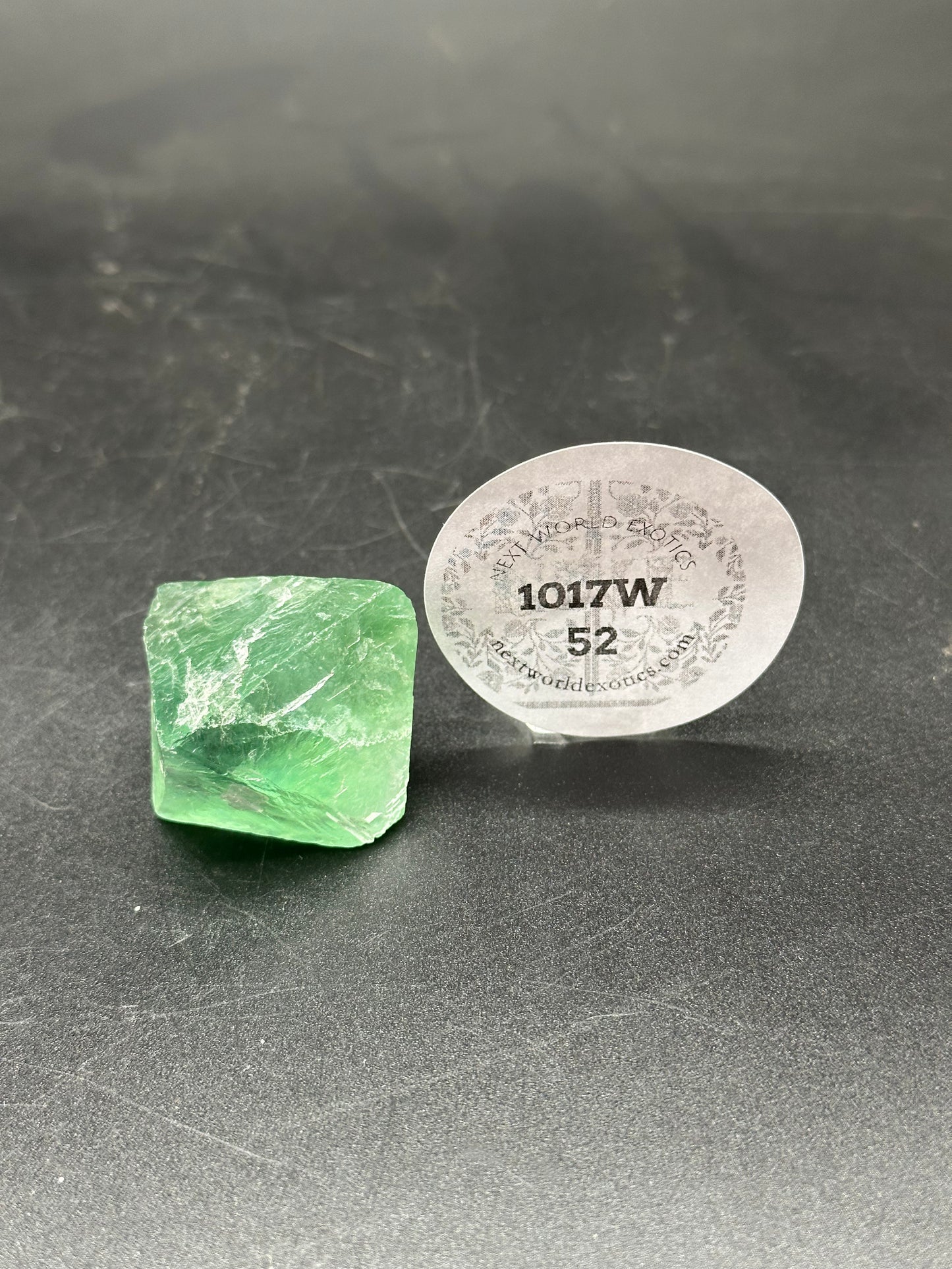 Fluorite Octahedron (1017W52) displayed on a black surface next to a round white sticker with text.