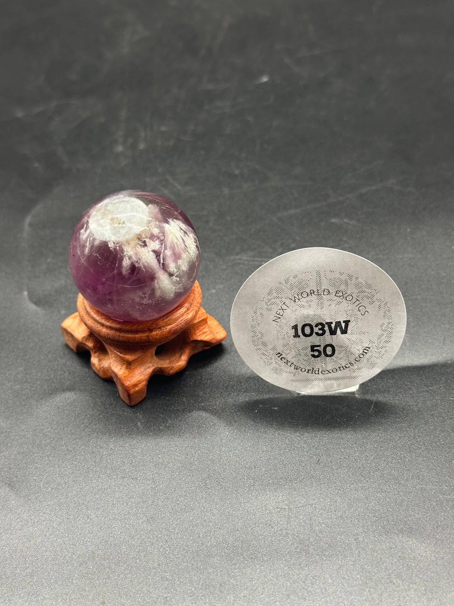 Fluorite Sphere (103W50) displayed on a wooden stand next to a round white object with text and numbers.