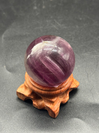 Fluorite Sphere (103W50) displayed on a wooden stand, highlighting its polished surface and spherical form in a still life setting.