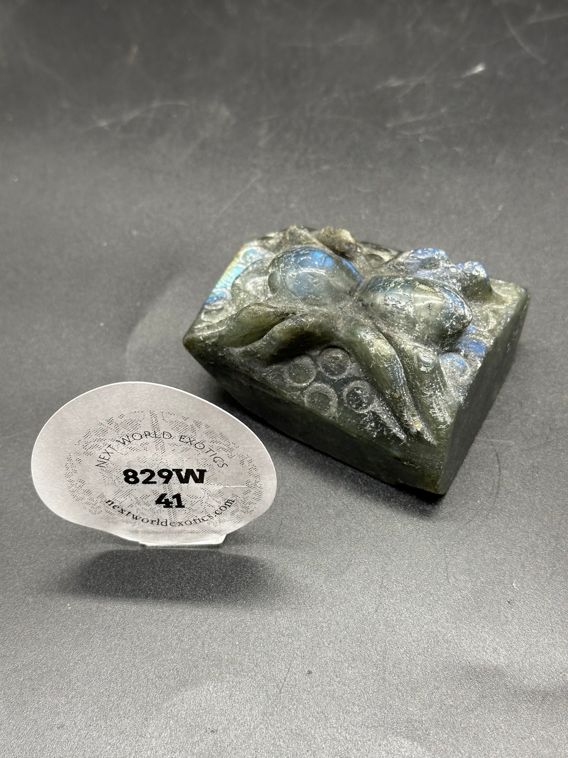 Labradorite Spider Carving (829W41) - A small square labradorite sculpture intricately carved into a spider, accompanied by a round white tag showing numbers.