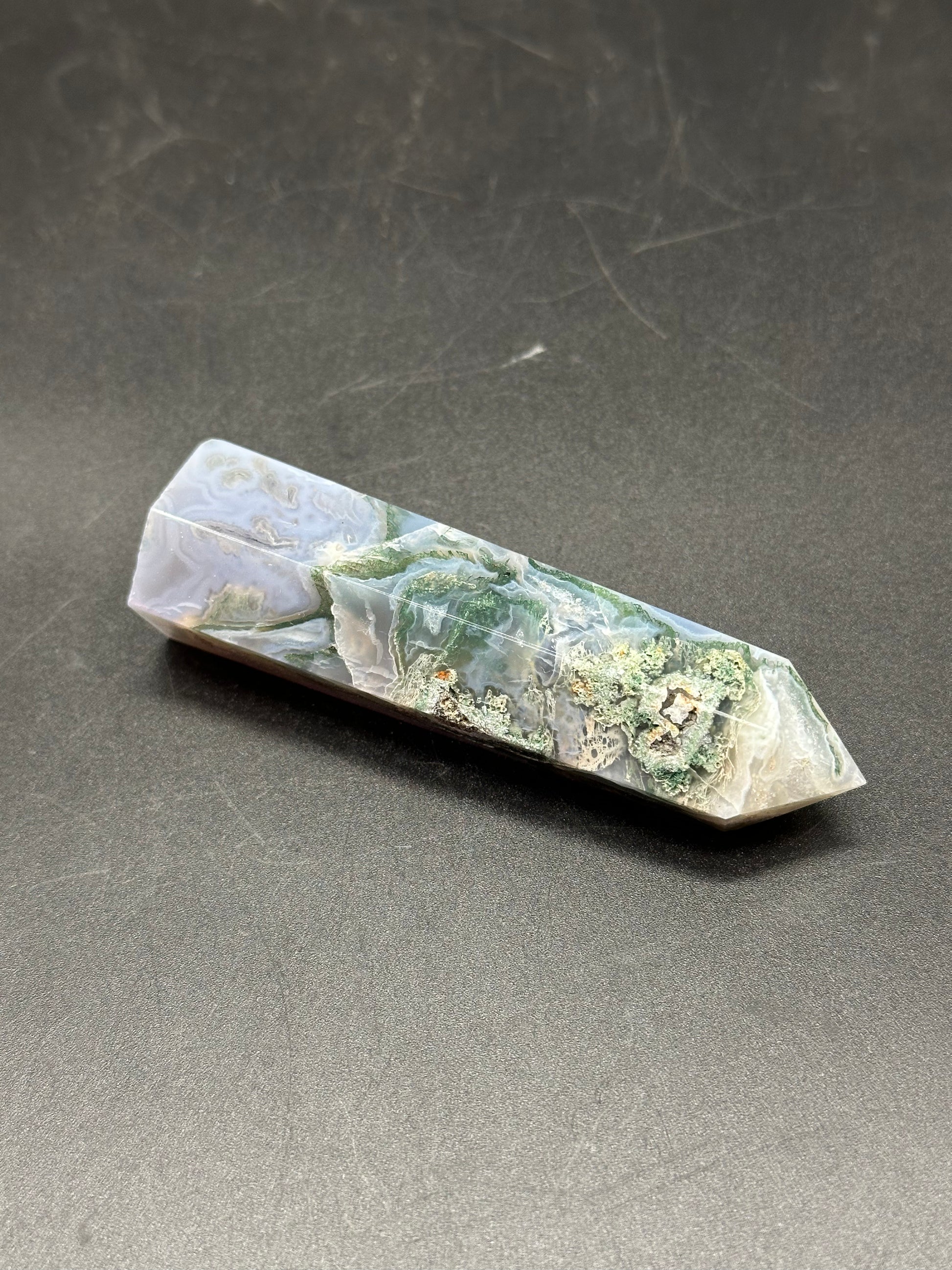 Moss Agate Tower (1017W43) crystal displayed on a textured surface, showcasing intricate patterns and natural formations.