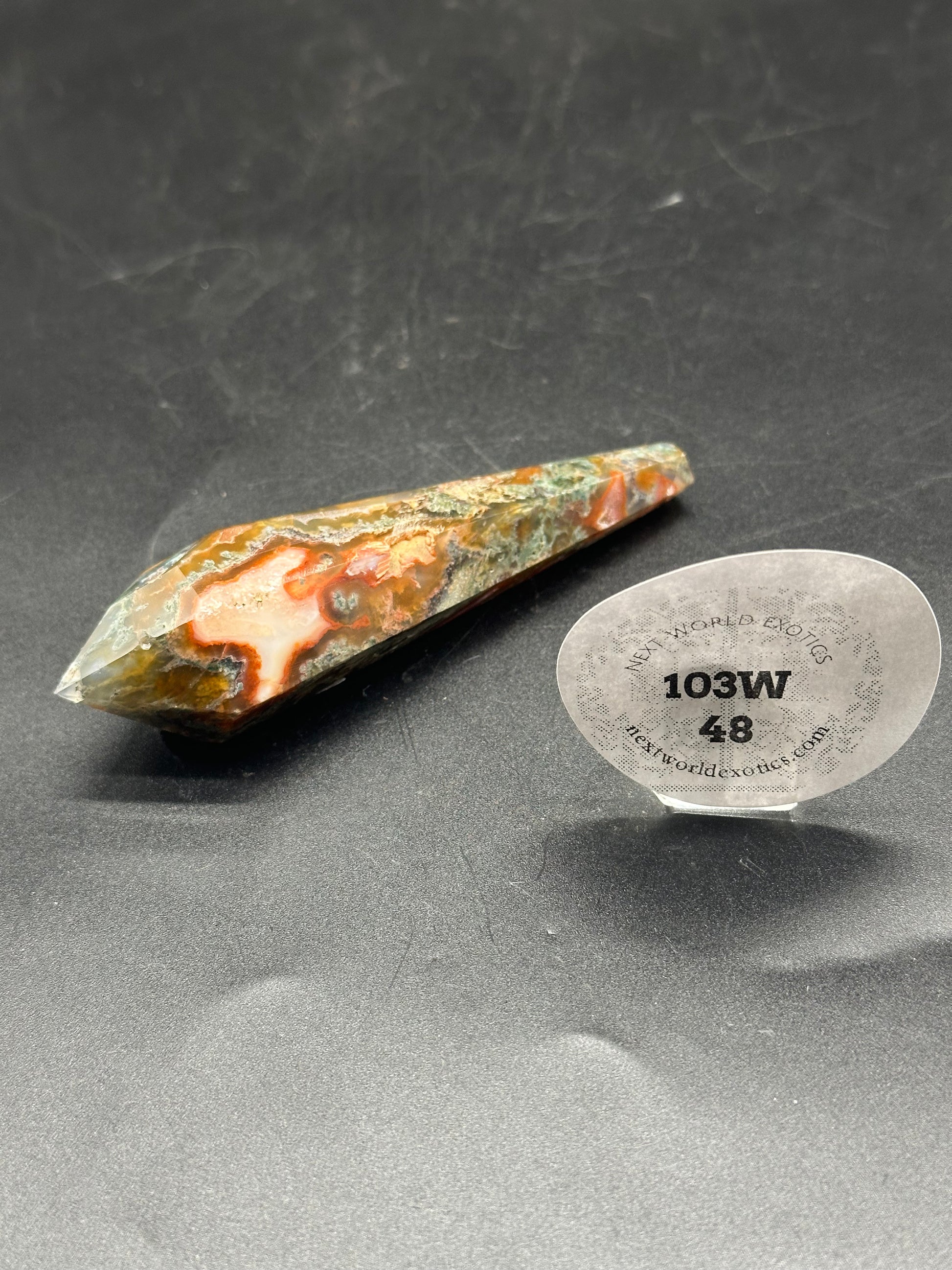 Red Moss Agate Scepter (103W48) featuring a pointed stone tip and a sticker with numbers and words on a black surface.