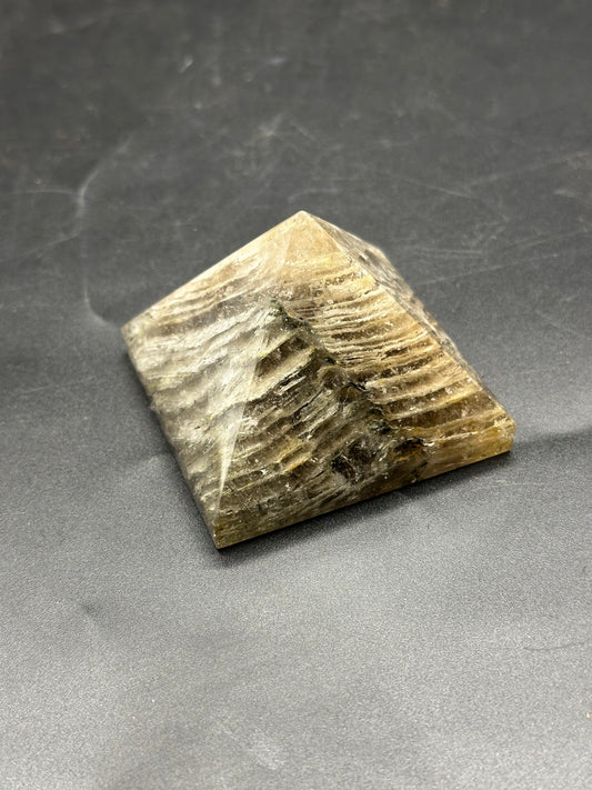 Thousand Layer Quartz Pyramid (1126W35) on a grey surface, showcasing its intricate pyramid shape and stone-like texture.