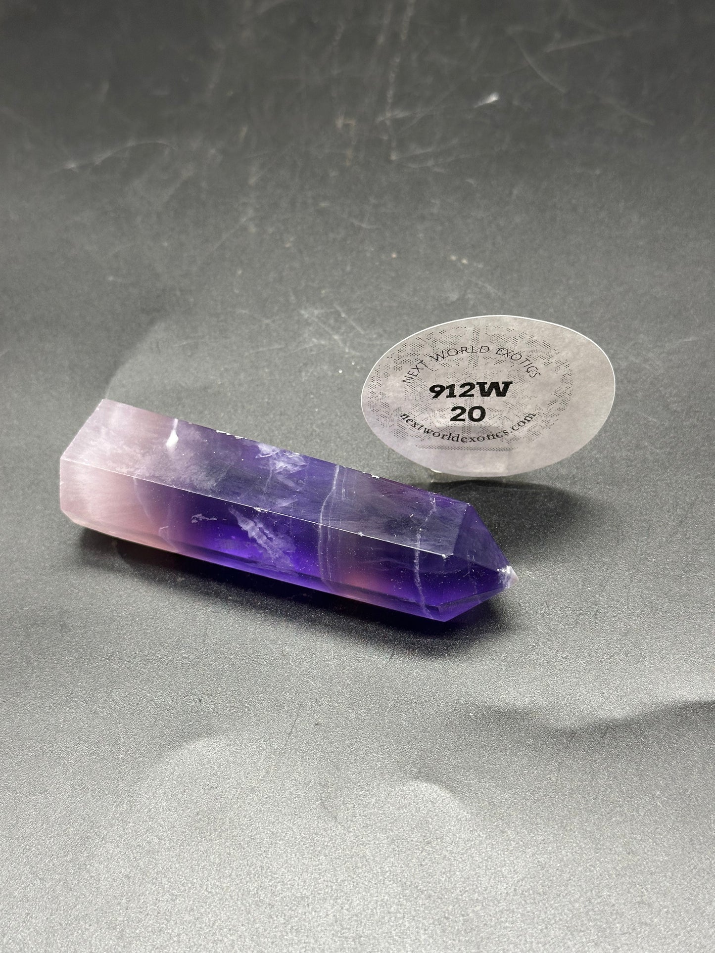 Violet Fluorite Tower (912W20) is a polished, pointed crystal with distinctive purple and white bands, displayed upright on a dark surface.